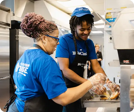 Two White Castle employees smile while preparing sliders together at a restaurant sandwich station.