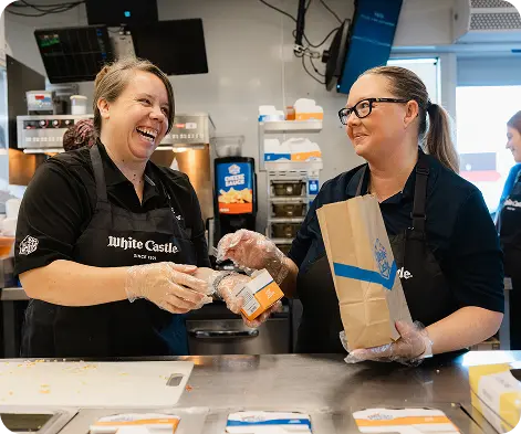 Two White Castle employees smile while packing a takeout order behind the restaurant counter.