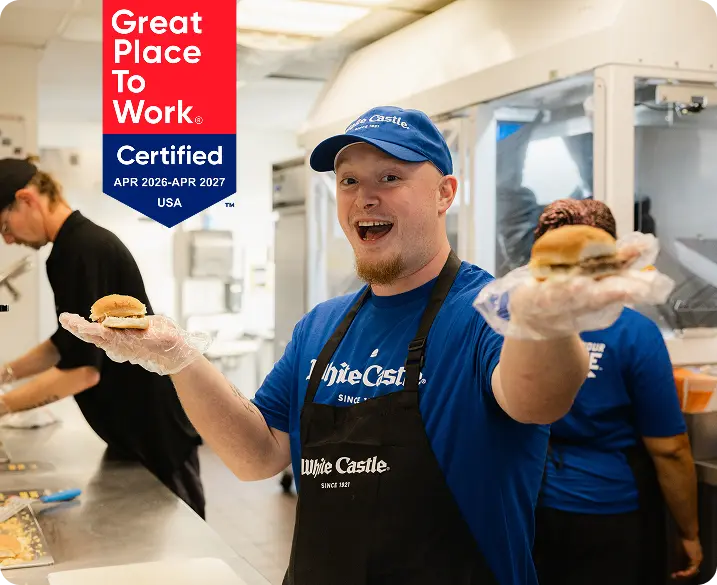 White Castle employee smiling and holding two sliders in a bright restaurant kitchen while coworkers prepare food.