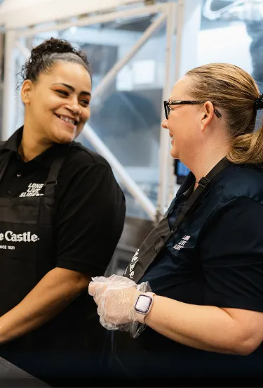 Two White Castle team members in aprons smile and chat together behind the restaurant counter.