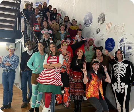 Employees in colorful costumes pose together on a staircase during a workplace Halloween celebration.