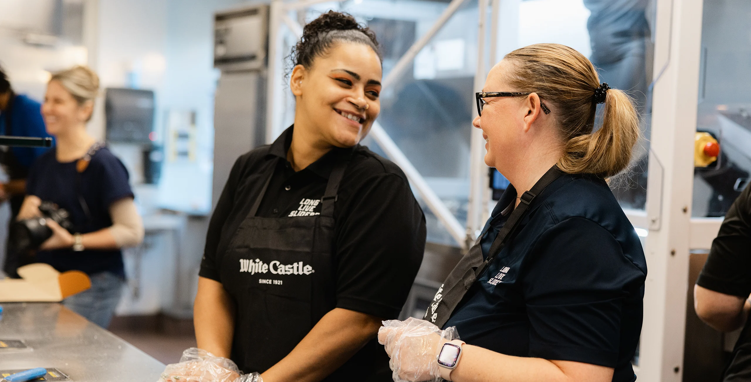 Two White Castle team members in aprons smile and chat together behind the restaurant counter.
