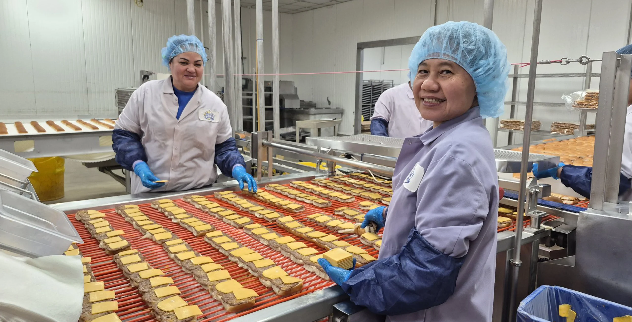 White Castle team members in white & blue uniform place cheese on sliders along a conveyor line in a food processing facility.
